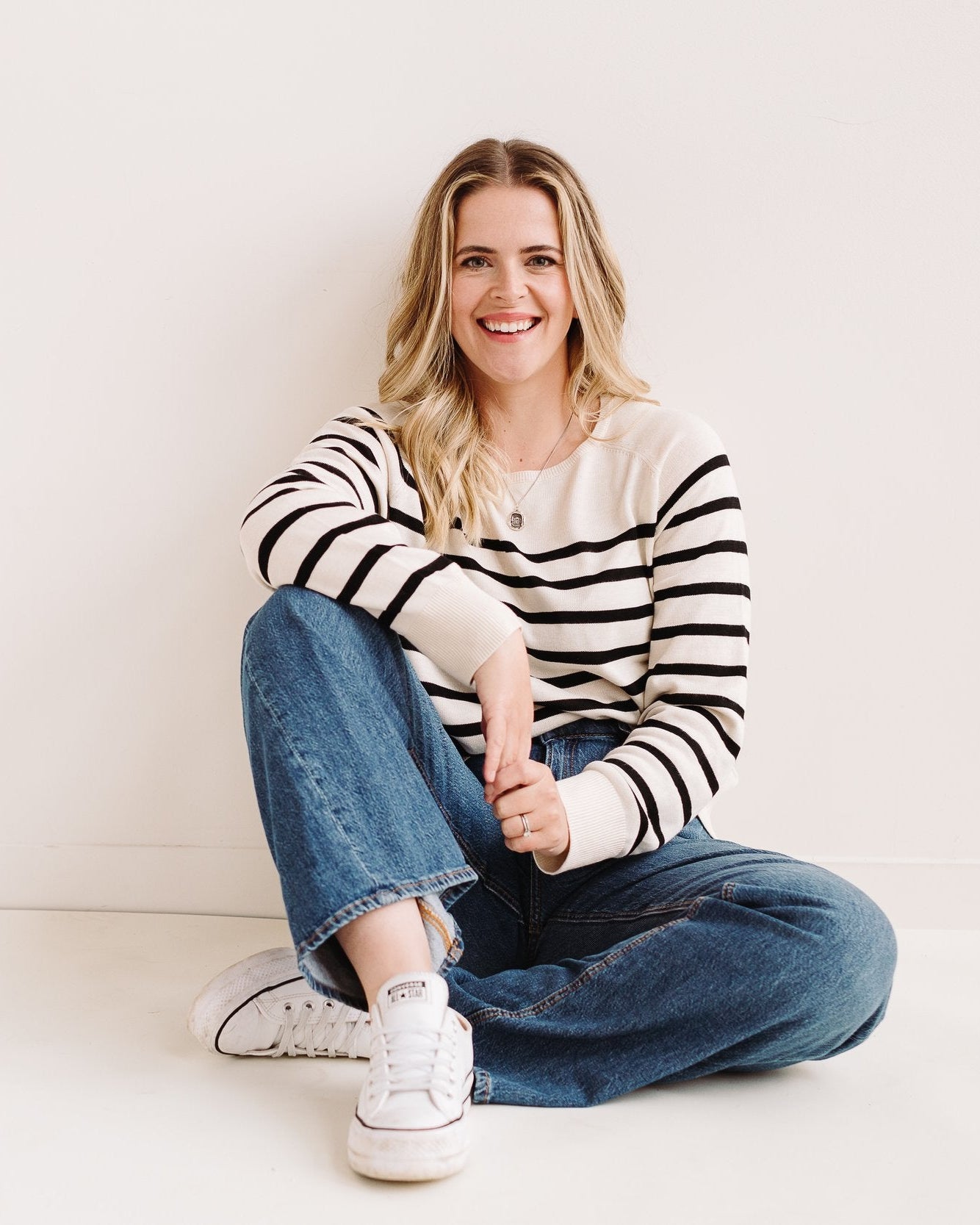 Woman sitting on the floor wearing a striped sweater and jeans against a white background.